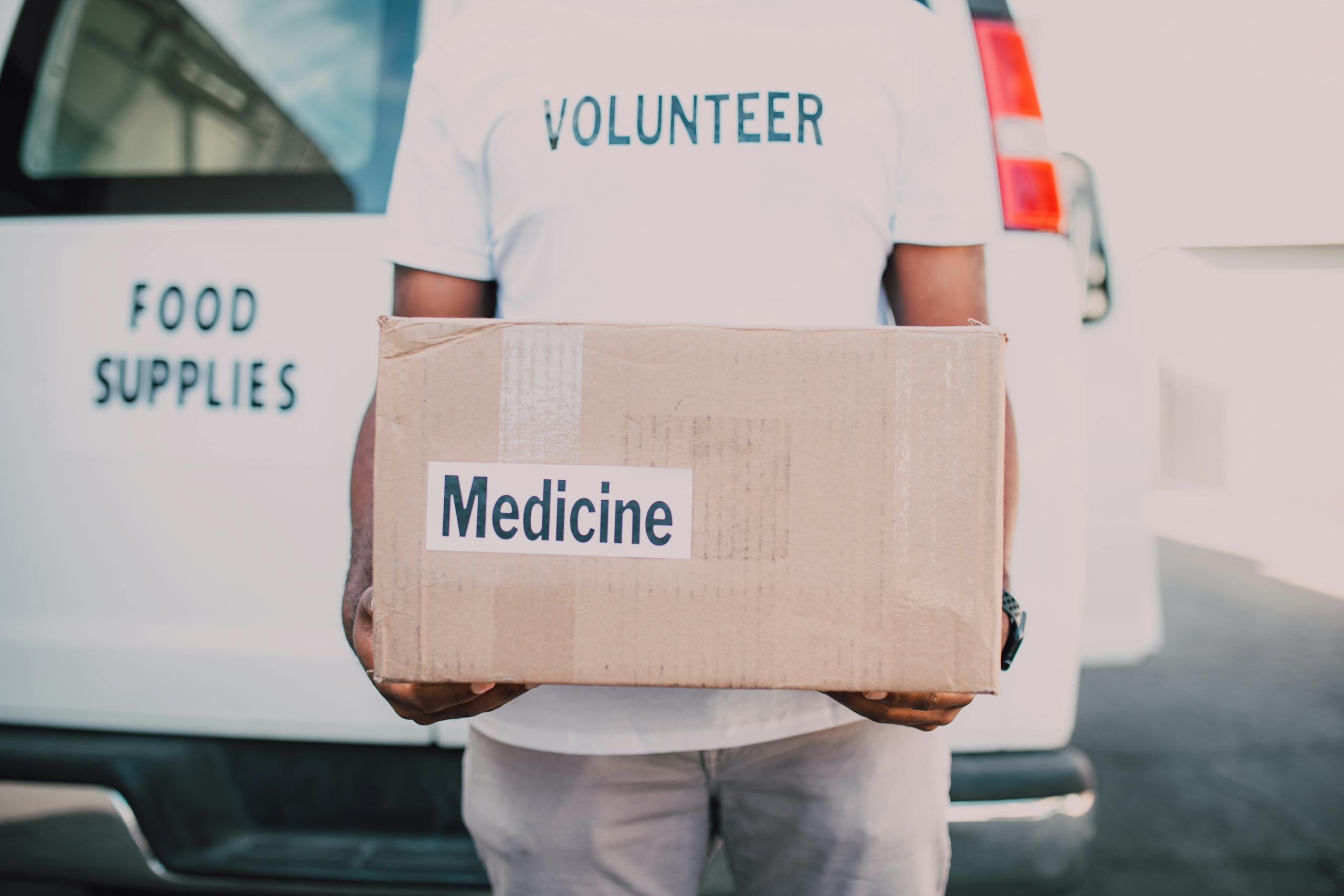 A volunteer holding a box labeled medicine near a delivery van marked food supplies.