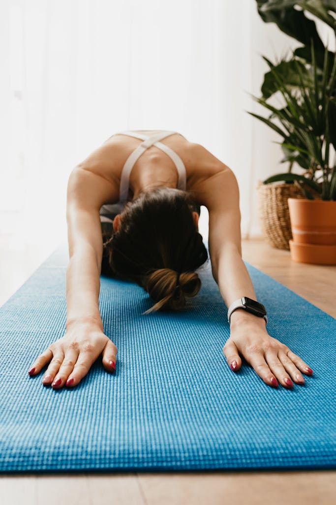 A woman practicing yoga in child’s pose on a blue mat indoors, promoting wellness and relaxation.